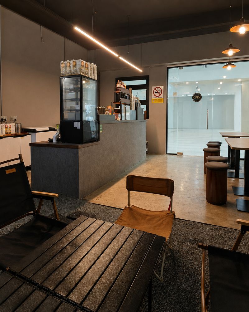 Inside Charming Cafe — the order counter and pastry case under warm pendant lights, with camping chairs in the foreground